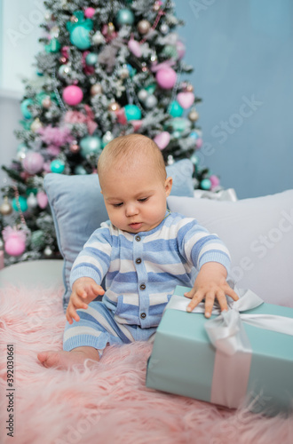baby boy in pajamas sitting on a fur blanket with pillows and gifts on the background of a Christmas tree
