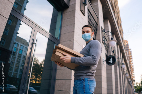 Low-angle shot of delivery man wearing medical mask ringing door for delivery carton boxes with hot pizza, looking at camera. Deliveryman in protective mask holding box with food.