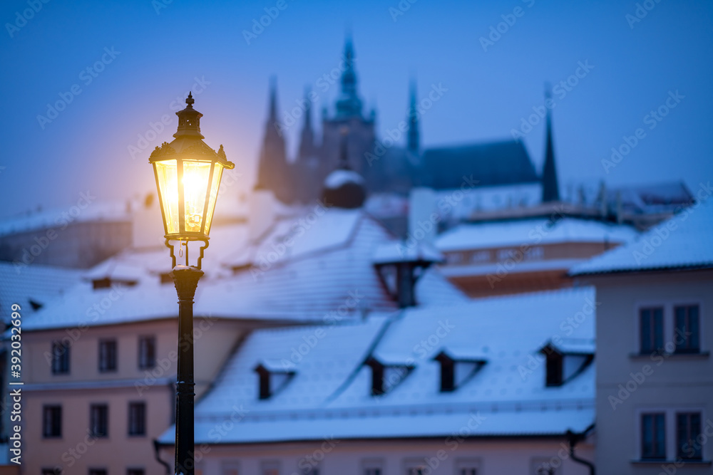 Φωτογραφία Romantic scene from winter Prague, with the lit lamp during dusk and snow covered roofs in the background, together with the silhouette of the Prague Castle at the horizon