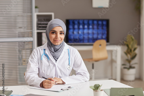 Portrait of young female doctor in white coat looking at camera while sitting at the table at office