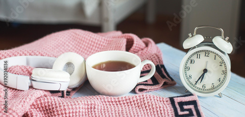 A cup of hot tea, knitted sweater, alarm clock and headphones in the bedroom. winter morning concept. banner