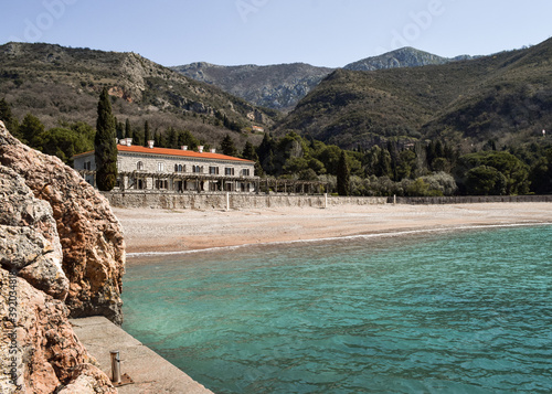 Rocks, blue sea coming onto the gravel beach, mountains and a two storey building. Queen's Beach, Montenegro.