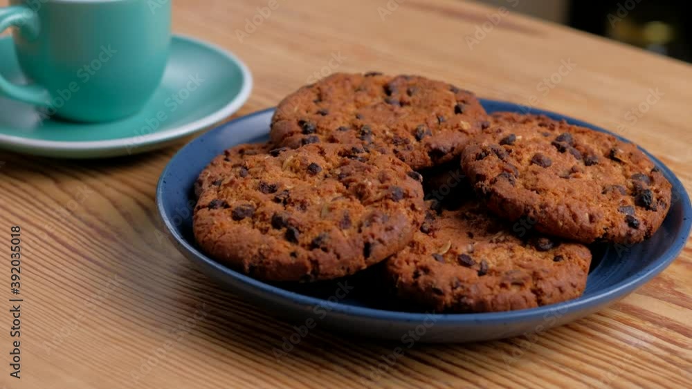 Appetizing homemade oatmeal cookies with piece of chocolate on blue plate with green cup of coffee on wooden table. Selective focus. Concept of Breakfast or dessert. Appetizing homemade oatmeal cookies with piece of chocolate on blue plate with green cup of coffee on wooden table. Selective focus. Concept of Breakfast or dessert.