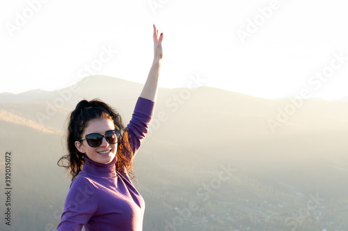 Young happy woman with raised up hand enjoying warm sunset evening in summer mountains.