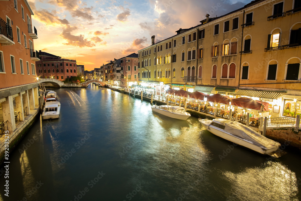Night scene of illuminated old buildings, floating boats and reflections in canal water in Venice, Italy.