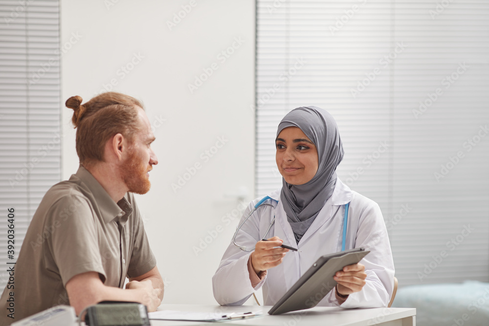 Female doctor in hijab using digital tablet while discussing methods of treatment with her patient at the table at hospital Female doctor in hijab using digital tablet while discussing methods of treatment with her patient at the table at hospital