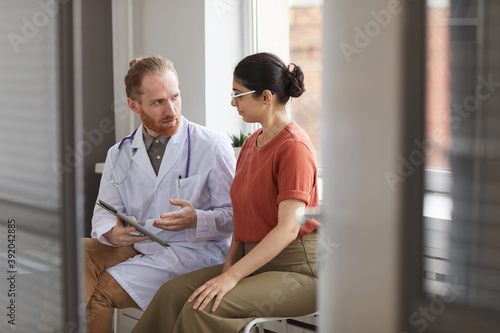 Male doctor in white coat talking to his patient while they sitting at the corridor of hospital