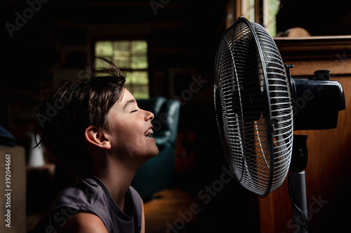 Young boy with eyes closed cooling off in front of a fan in dark room.
