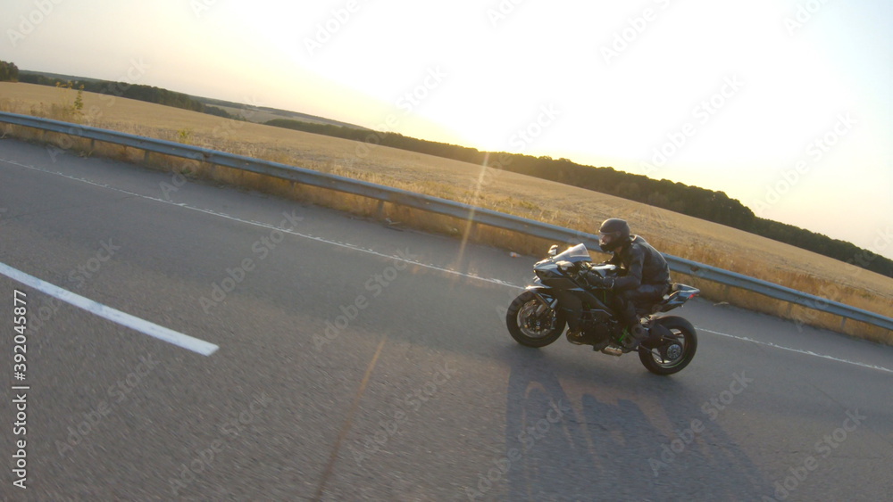 Fototapeta premium Camera moving around of biker riding on modern sport motorbike at highway. Motorcyclist racing his motorcycle on country road at sunset. Man driving bike during trip. Concept of freedom and adventure