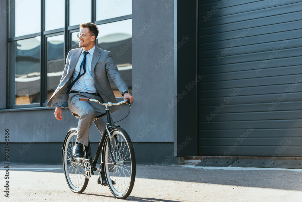 Positive businessman in formal wear and eyeglasses riding bicycle near building Positive businessman in formal wear and eyeglasses riding bicycle near building