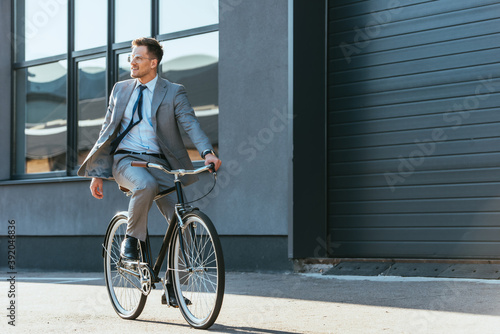 Fototapeta Naklejka Na Ścianę i Meble -  Positive businessman in formal wear and eyeglasses riding bicycle near building