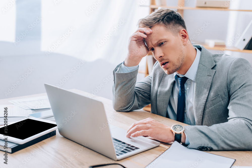 Thoughtful businessman using laptop near papers and digital tablet on blurred foreground Thoughtful businessman using laptop near papers and digital tablet on blurred foreground