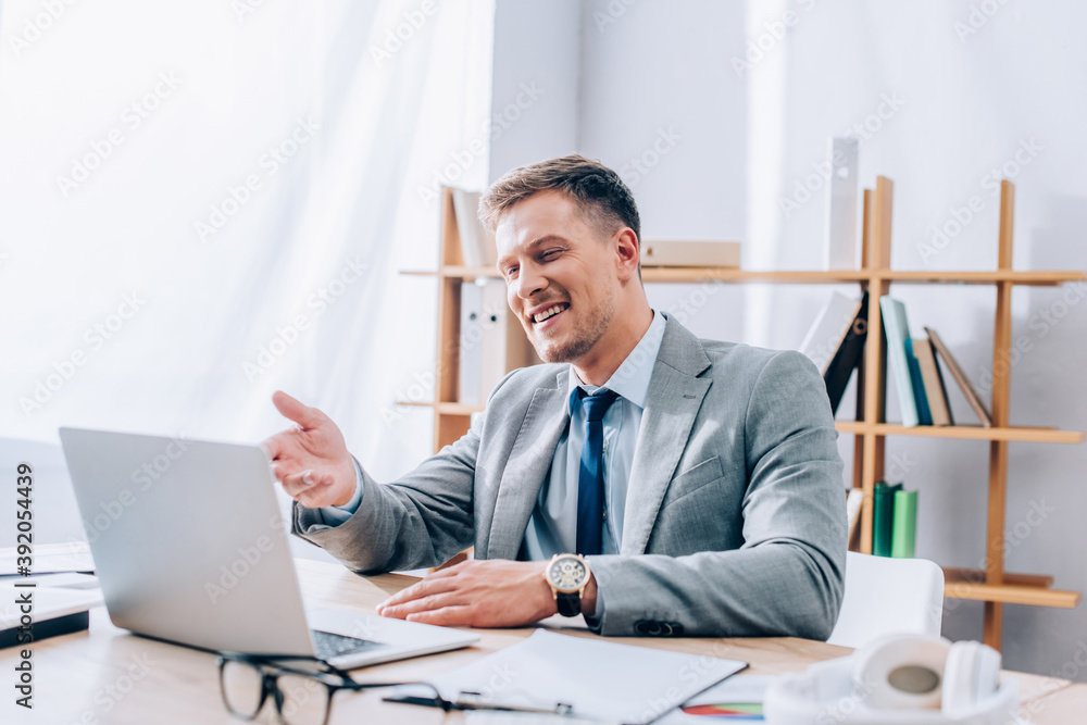 Smiling businessman having video chat on laptop near headphones and papers on blurred foreground Smiling businessman having video chat on laptop near headphones and papers on blurred foreground