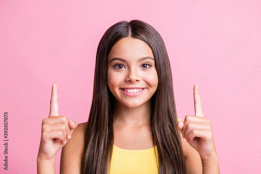 Photo of trendy brown long haired happy girl point finger empty space wear yellow singlet on pink color background Photo of trendy brown long haired happy girl point finger empty space wear yellow singlet on pink color background