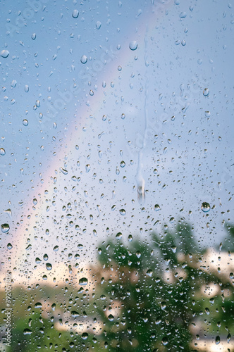 The focus is on the raindrops on the glass surface of the window, with rainbows and green foliage on the background