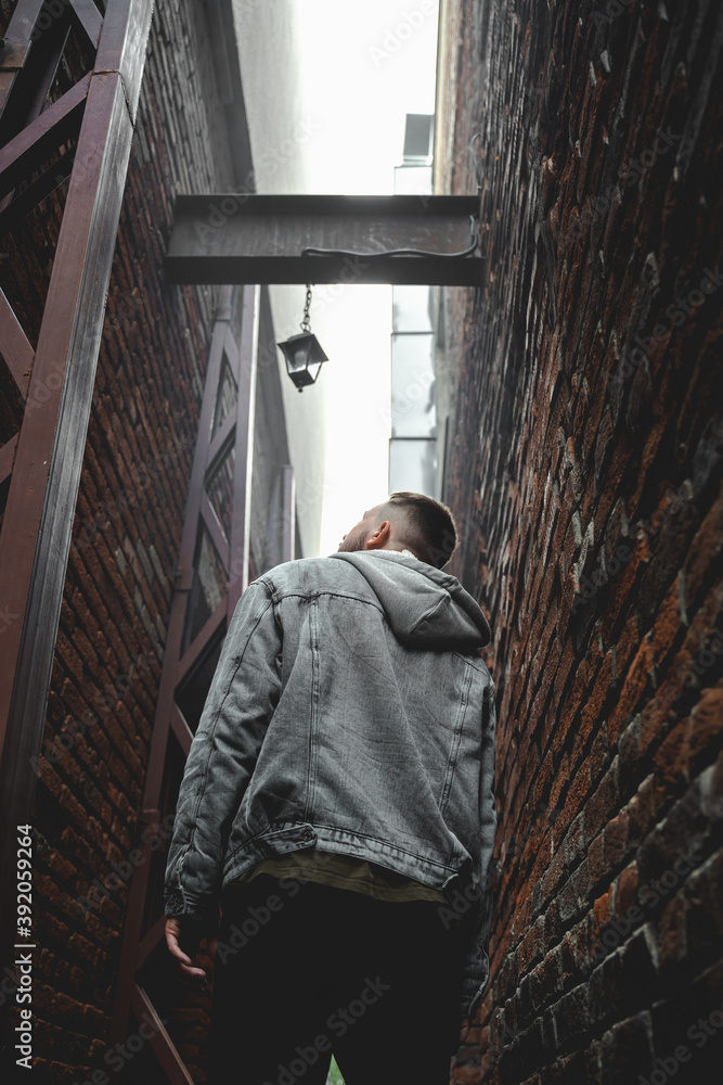 Young bearded man in denim jacket walks through brick alley and looks up