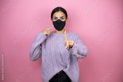 Beautiful woman wearing a casual violet sweater over pink background pointing the mask. Warning expression with negative and serious gesture on the face.