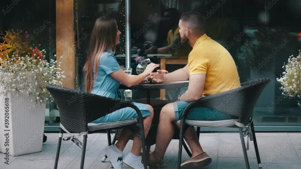 Romantic date of young happy couple in love smiling talking sitting at table in cafe outside on summer day, medium view in slow motion. Cute guy and girl spend leisure time in restaurant holding hand. Romantic date of young happy couple in love smiling talking sitting at table in cafe outside on summer day, medium view in slow motion. Cute guy and girl spend leisure time in restaurant holding hand.