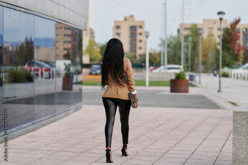 Back view of a black woman wearing a brown suit holding a laptop and a container of coffee walking next to a building. business concept