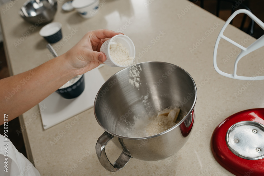 A close photo of the hand of a young woman who is putting an almond flour from the cup into the stainless steel bowl of the stand mixer.