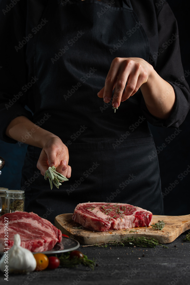 Close-up view of chef in black uniform pours rosemary to raw steak on wooden chopped board. Backstage of preparing grilled pork meat at restaurant kitchen on dark blue background. Frozen motion.
