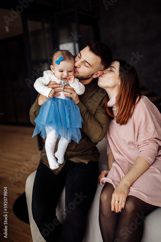 Loving mother and caring father with cute toddler sit on the sofa near decorated Christmas tree, happy dad gently kiss little daughter, smiling, winter holidays at home concept