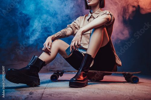 Dressed in trendy and simple clothing girl with short haircut and tattoo poses sitting on skate and holding smartphone in smokey background.