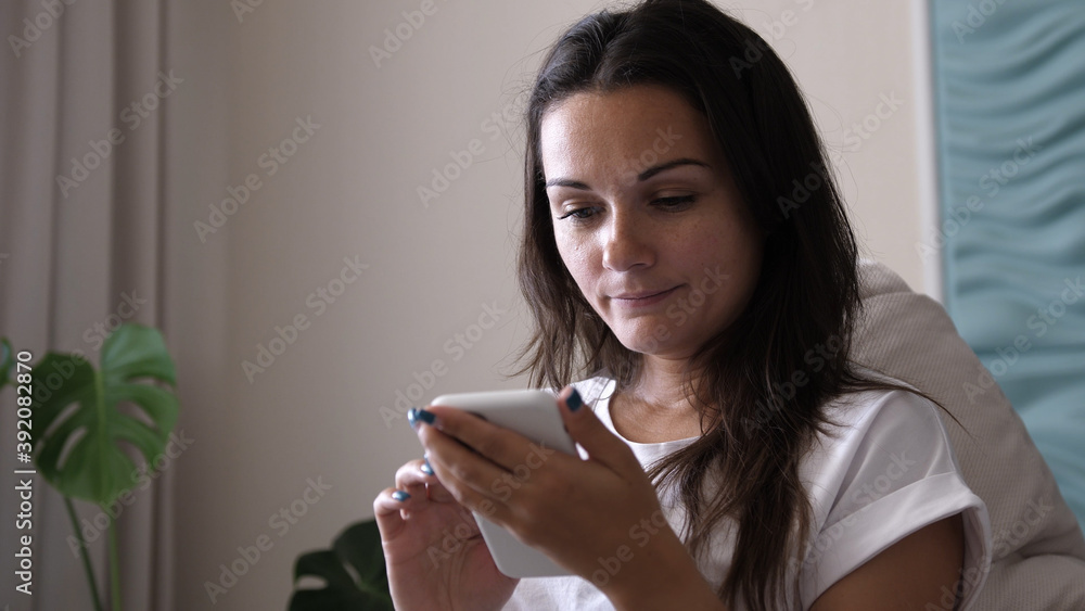 Happy young woman holding smart phone looking at cellphone screen enjoying using mobile apps for shopping having fun playing games chatting in social media sit on couch at home Happy young woman holding smart phone looking at cellphone screen enjoying using mobile apps for shopping having fun playing games chatting in social media sit on couch at home