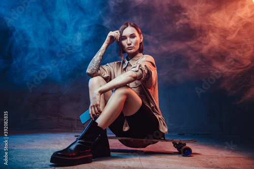 Dressed in stylish casual clothing girl with modern smartphone and tattoo on her right hand sits on skate in smokey background.