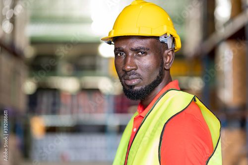 Close up of African American male warehouse worker in safety vest and helmet working in in industry storage warehouse with shelf pallet product or spare parts parcel blur background © amorn