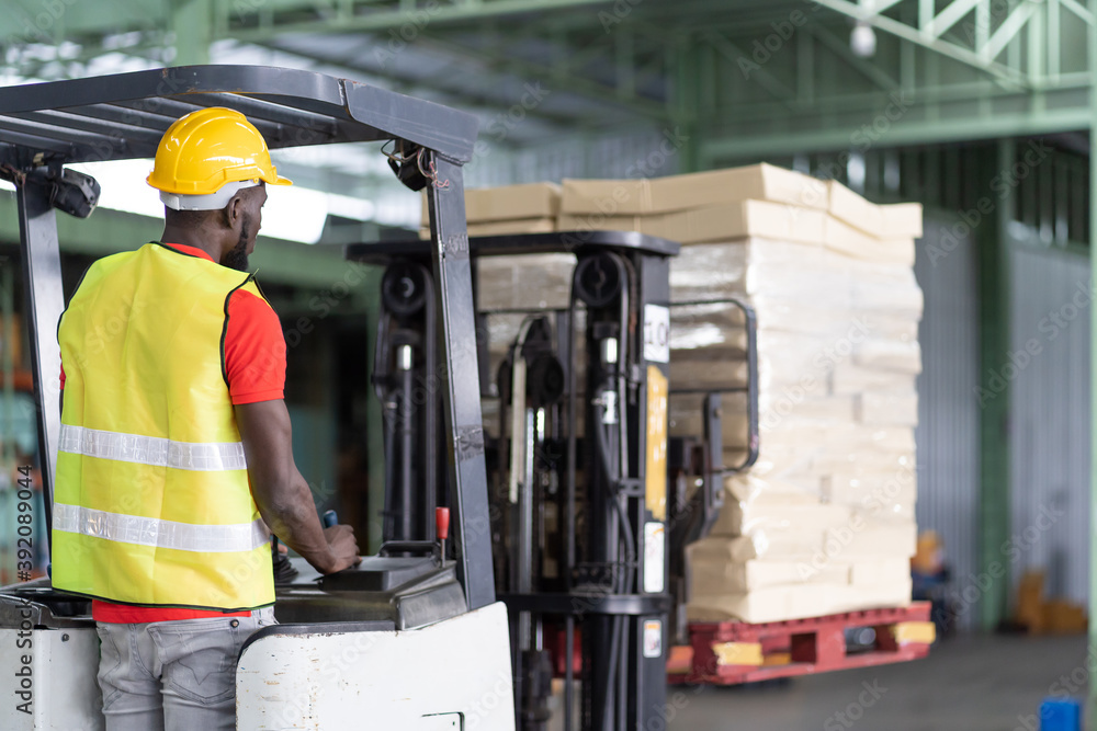 Fototapeta premium African American male warehouse worker in safety vest and helmet driving and operating on forklift truck for transfer products or parcel goods in the industrial storage warehouse. multicultural worker