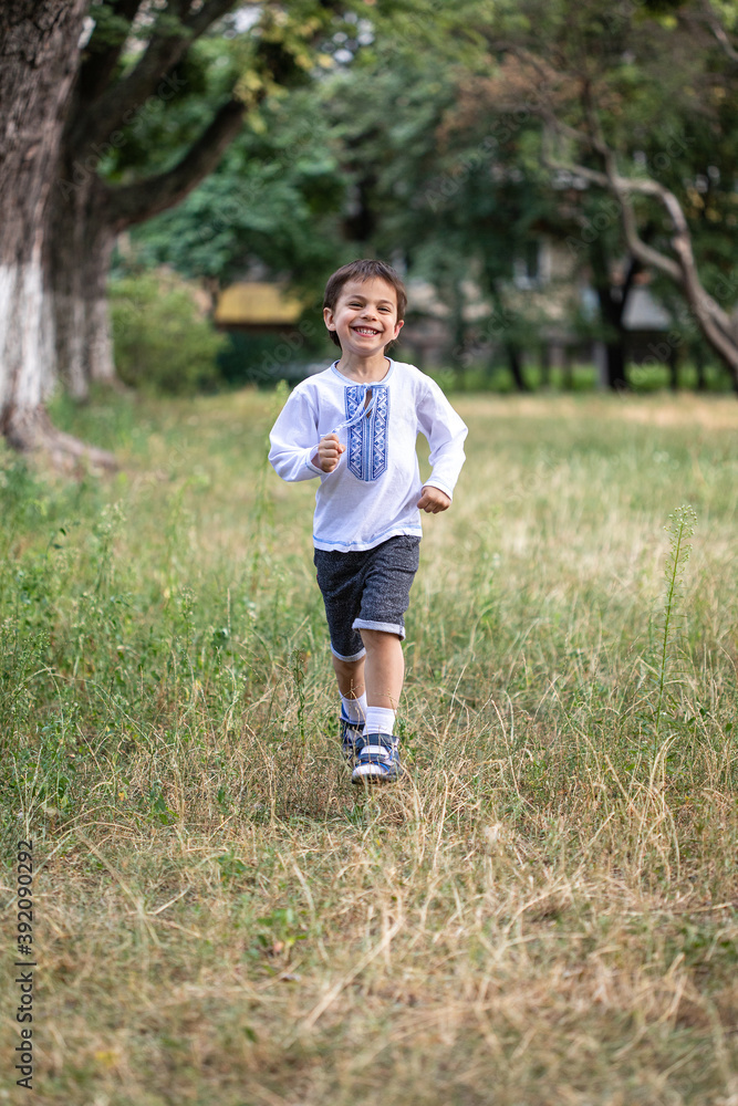 Little adorable boy in shirt with Ukrainian ornament running in park and smiling Little adorable boy in shirt with Ukrainian ornament running in park and smiling