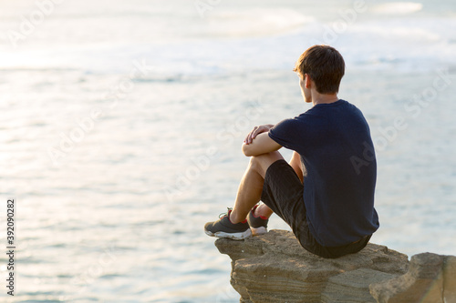 Thoughtful young man sitting alone on top of a cliff above the ocean view at sunset. photo