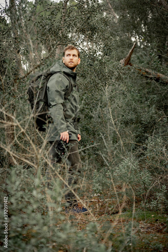 Young mountaineer man dressed in dark green in the undergrowth looking behind with photo camera, dark and desaturated style, adventure concept.