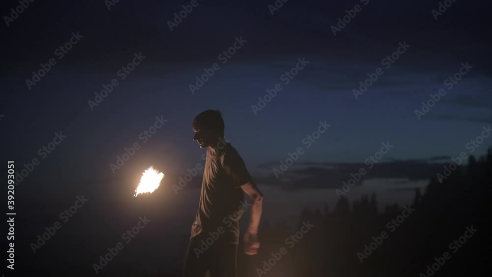 Graceful male fireshow performer making fire dance with two metal fans lit with flames on night mountains meadow Graceful male fireshow performer making fire dance with two metal fans lit with flames on night mountains meadow