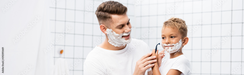 Smiling father looking at son with shaving foam on face holding safety razor near cheek in bathroom, banner