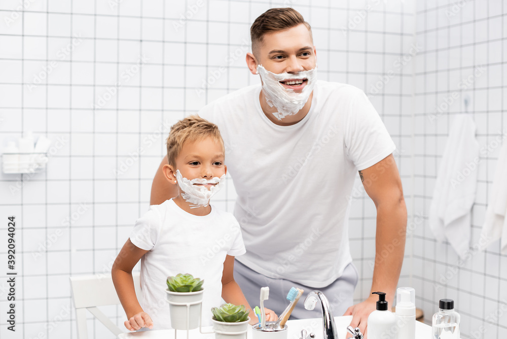 Smiling father and son with shaving foam on faces standing near sink in bathroom