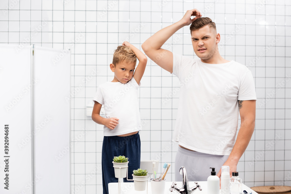 Father and son with armpits smelling badly looking at camera, while standing near sink with toiletries in bathroom Father and son with armpits smelling badly looking at camera, while standing near sink with toiletries in bathroom