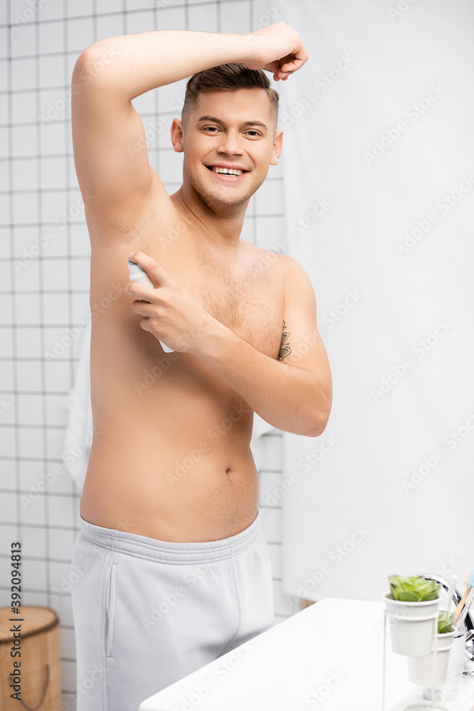 Obraz premium Cheerful shirtless man looking at camera while spraying antiperspirant in bathroom