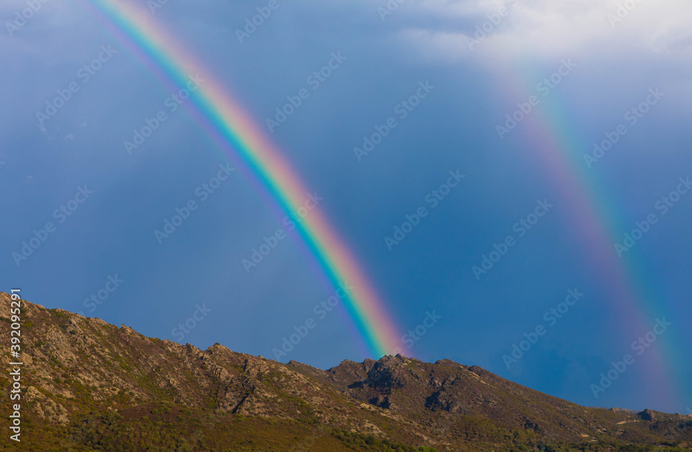 Panorama view of full double rainbow with blue sky and mountain range in background. Near Saint Florent, Corsica