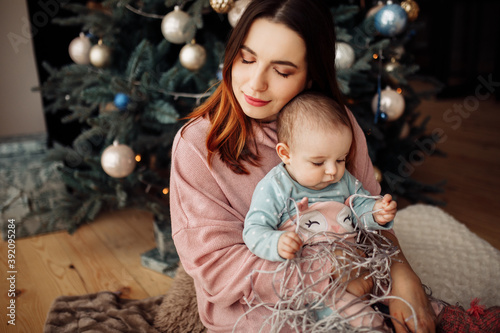 Pretty woman with lovely toddler sit near Christmas tree, tender mother hold cute daughter in arms, smiling, little baby girl look at the garland, winter holidays at home concept