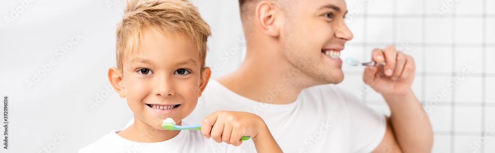 Smiling son with toothbrush looking at camera and standing near father looking away, while brushing teeth in bathroom, banner Smiling son with toothbrush looking at camera and standing near father looking away, while brushing teeth in bathroom, banner