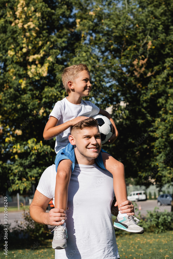 Happy son holding ball and riding piggyback on smiling father in park on blurred background Happy son holding ball and riding piggyback on smiling father in park on blurred background