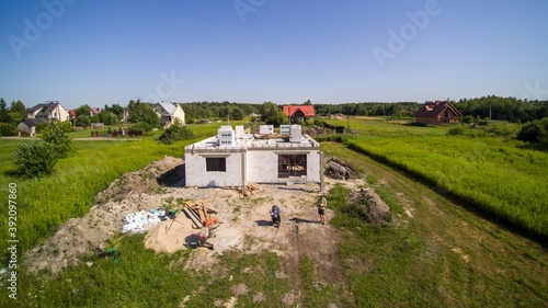 Aerial view on detached house under construction. House in basic state. Located on green plot in small village. Wooden roof structure. Meadows around.