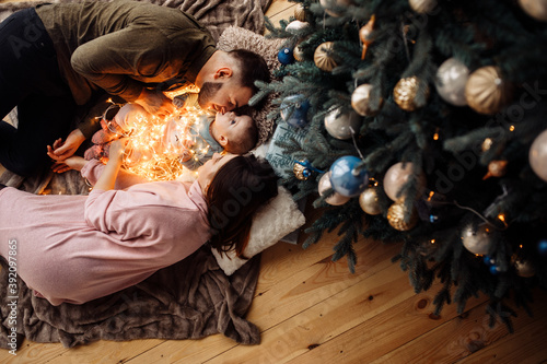 Caring parents lay with funny baby girl on blanket at home, loving mom and dad gently kiss little child at the cheeks, happy family spend winter holidays together, xmas concept