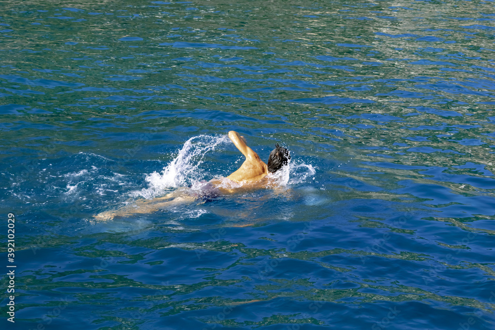 Fototapeta premium A man swims in a bright blue-green sea water. Naked male silhouette waving his hands, splashing water drops. Summer sports background with copy space