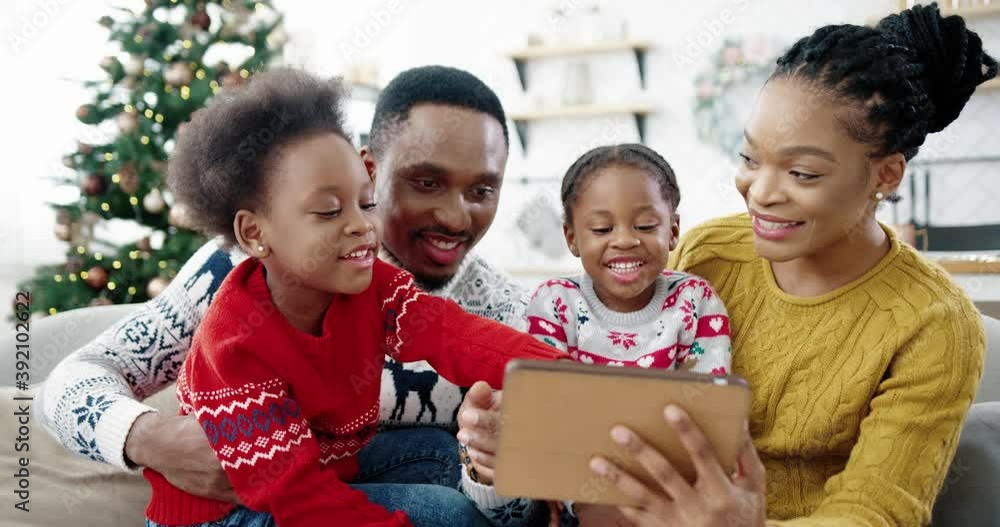 Close up of happy African American family at home sitting in decorated room near Christmas tree and using tablet Small cute kids sitting with mom and dad and tapping on tablet choosing presents online Close up of happy African American family at home sitting in decorated room near Christmas tree and using tablet Small cute kids sitting with mom and dad and tapping on tablet choosing presents online