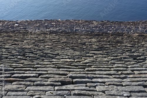 Abstract dam wall, looking down towards deep blue water, sunny stones