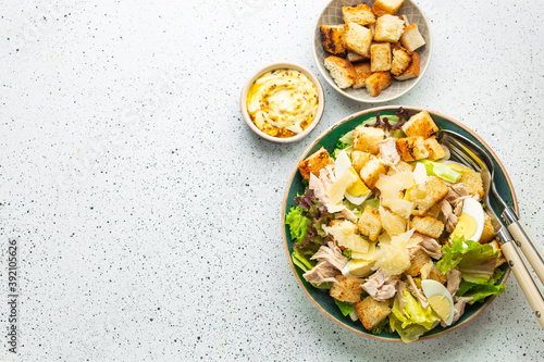 Fresh Caesar salad with lettuce salad, chicken breast, boiled eggs and croutons in ceramic bowl with dressing on the side on white table. Classic healthy salad, top view, space for text