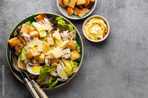 Fresh Caesar salad with lettuce salad, chicken breast, boiled eggs and croutons in ceramic bowl with dressing on the side on gray table. Classic healthy salad, top view, space for text